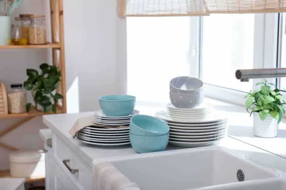Stacked dishes beside a kitchen sink and window in soft natural light