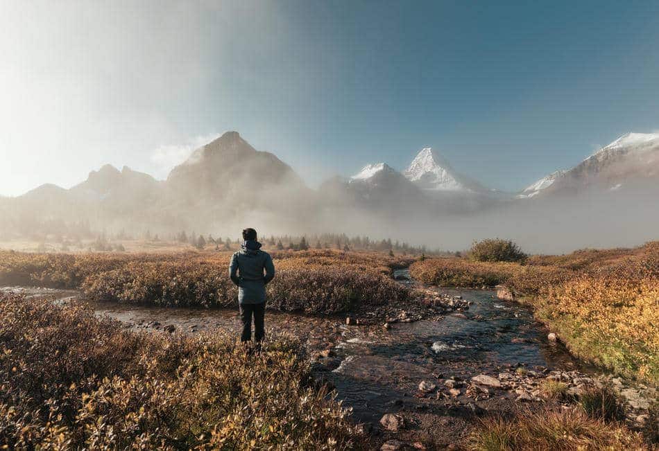 Person standing near a mountain stream looking toward a distant landscape.