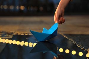 A hand gently releases a blue paper boat onto calm water, symbolizing letting go of control and moving forward with intention.