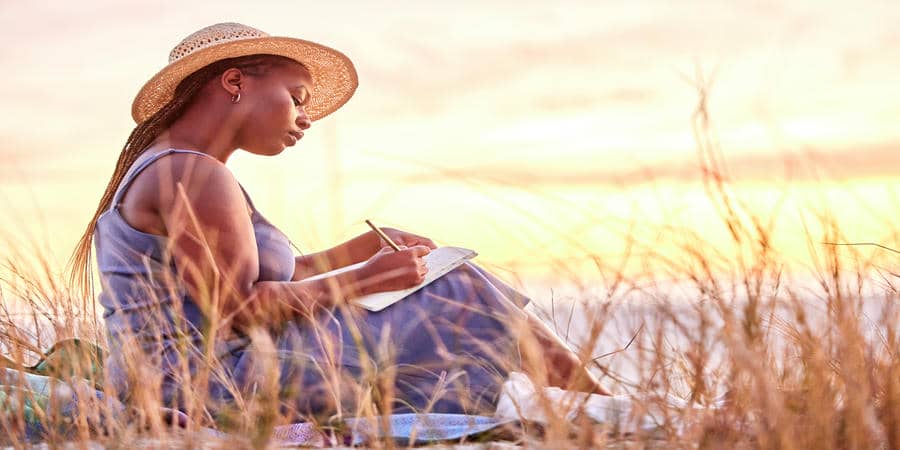 Person writing in a journal while sitting in tall grass at sunset, exploring thoughts and personal patterns through reflection.