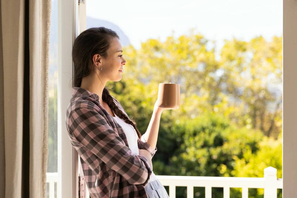 Woman holding a cup of coffee while looking through an open window, enjoying a peaceful morning moment.
