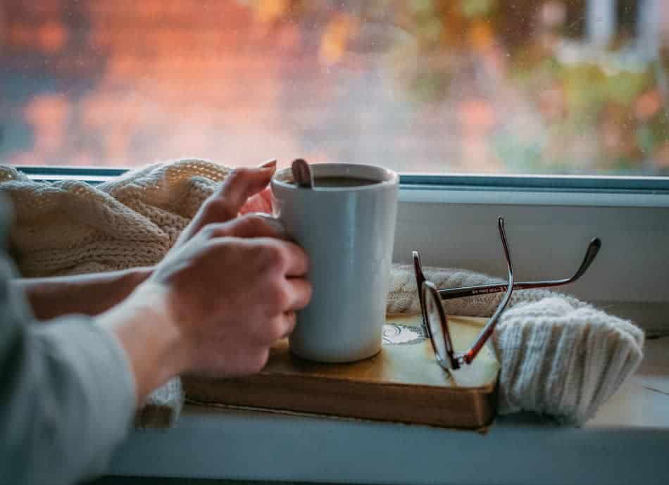 Hands holding a warm mug beside a window with a book and glasses nearby