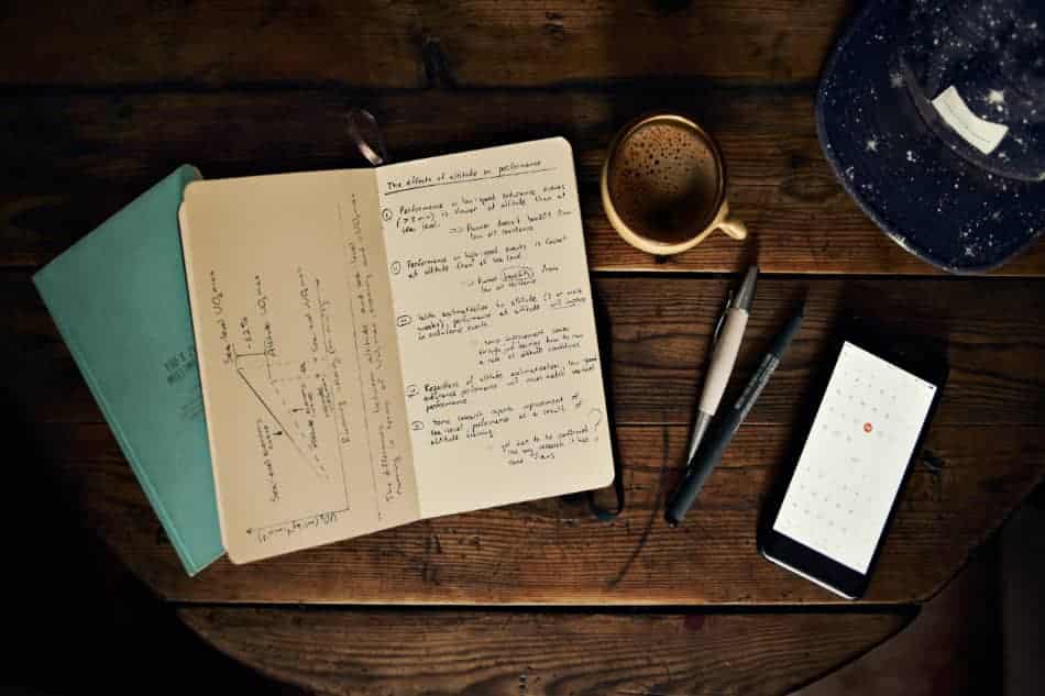 Open notebook, handwritten notes, phone, and writing tools scattered across a wooden table