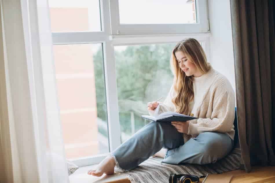 Young woman writing in a journal while sitting by a bright window, reflecting quietly and practicing self-awareness.