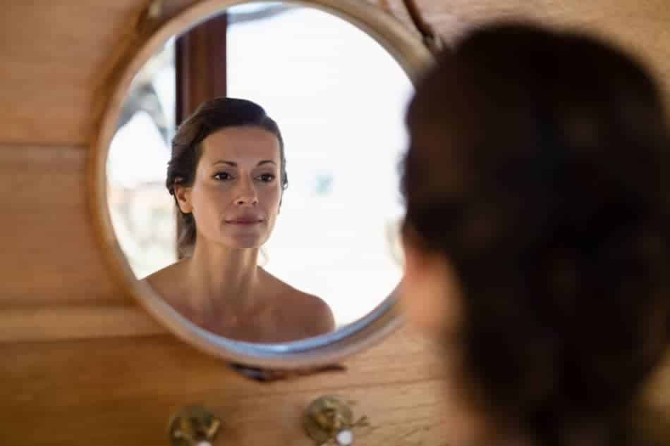 Woman quietly looking at her reflection in a small round mirror, symbolizing the first moment of self-awareness before self-judgment.