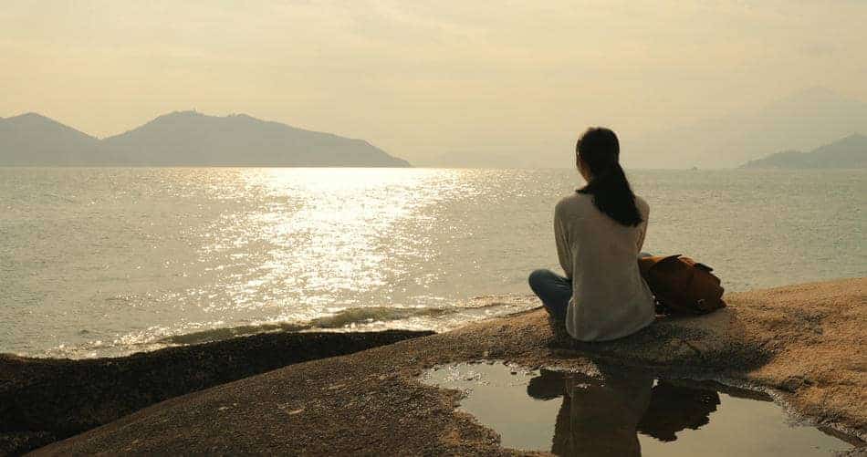Person sitting quietly by the water at sunset, reflecting in a calm and peaceful moment