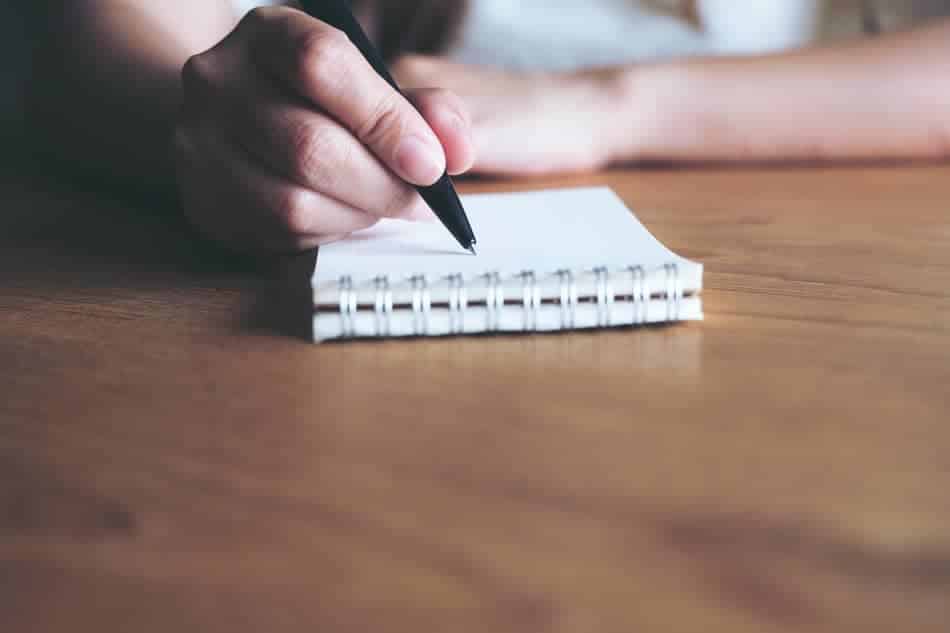 Close-up of a hand writing in a small notebook on a wooden desk, representing a simple first step