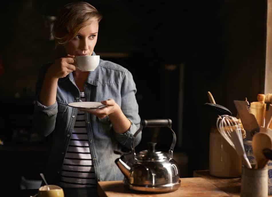 Woman standing quietly in a warm kitchen holding a cup of tea in soft window light
