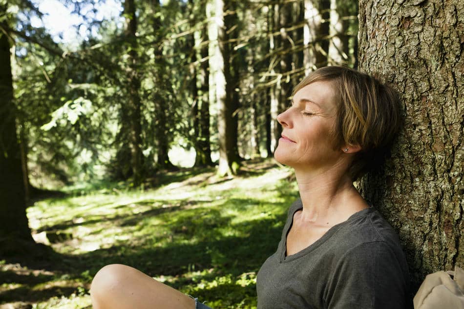 A woman rests against a tree with her eyes closed, reconnecting with herself through a quiet moment in nature.
