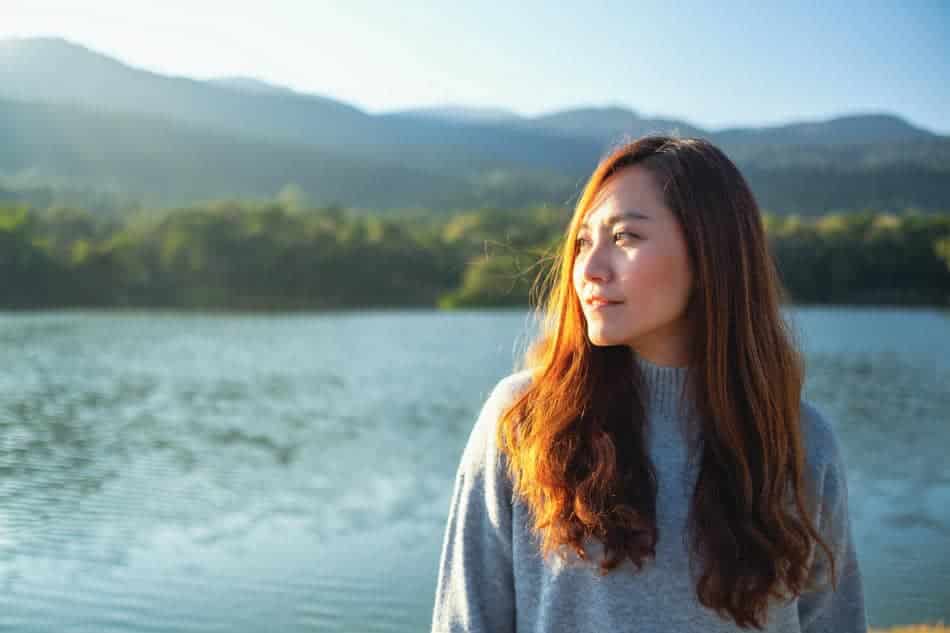 A woman stands beside a peaceful lake, looking toward the horizon with calm confidence and clarity.