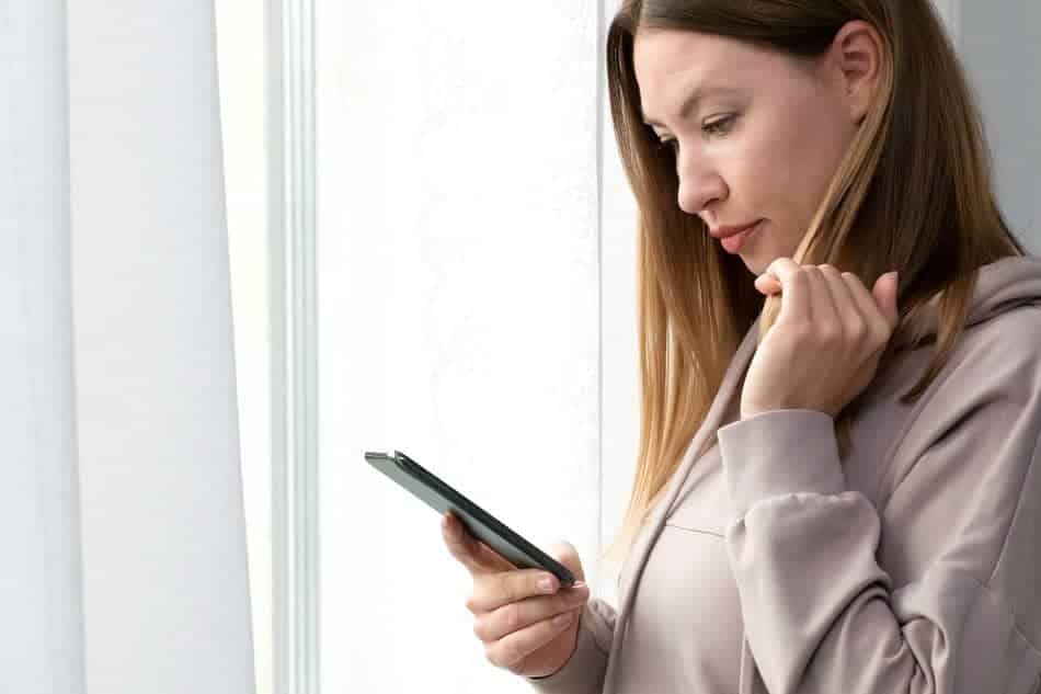 A woman thoughtfully reads a message on her phone by a window, pausing before responding.