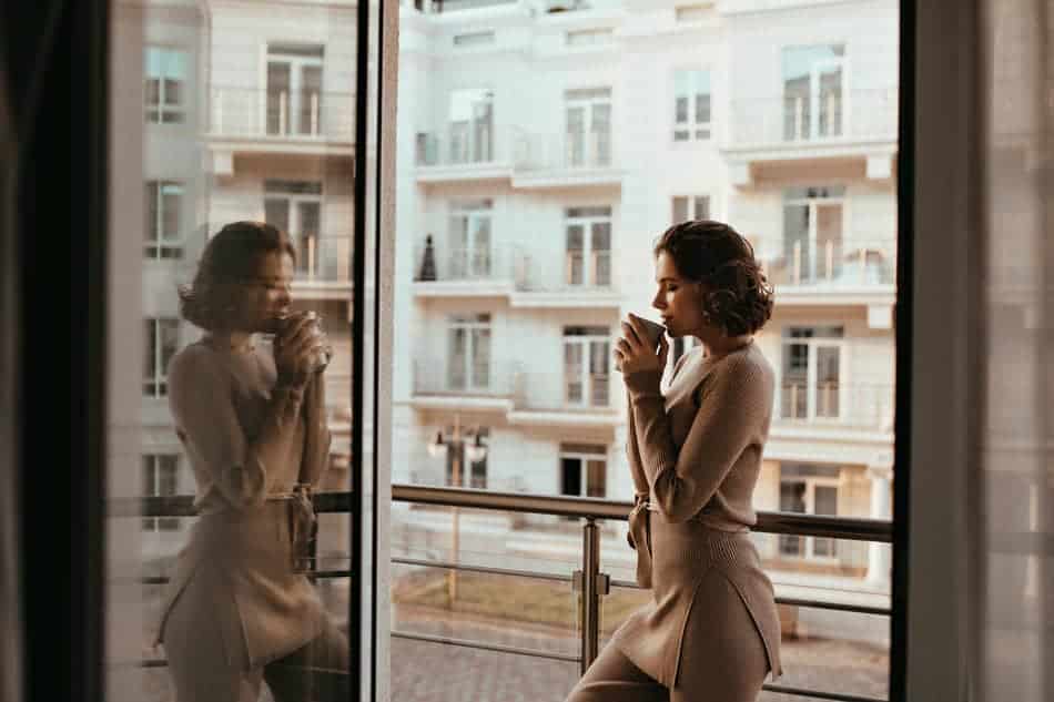 Woman holding a warm drink by a window, her reflection beside her as she pauses for quiet self-reflection.