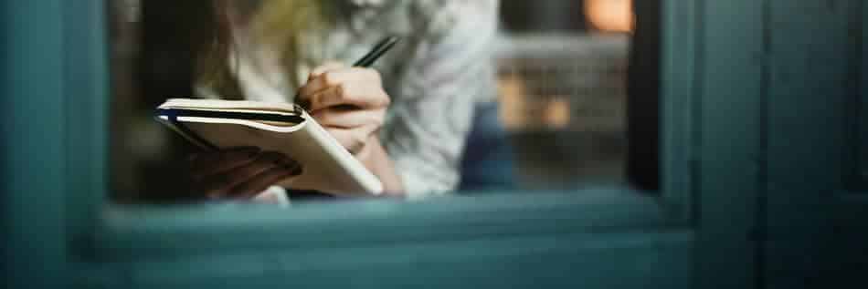 Woman writing in a journal beside a window, reflecting quietly and putting her thoughts into words.